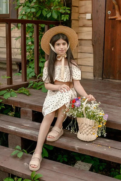 Souriant fille préadolescente aux cheveux bruns assis sur le seuil du chalet rural avec panier de fleurs sauvages — Photo