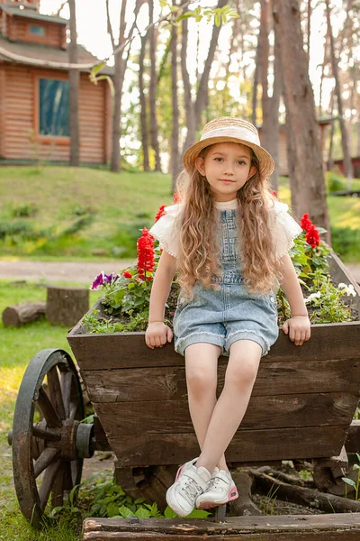 Dromerig mooi meisje zitten op vintage houten kar versierd als bloembed op landgoed — Stockfoto
