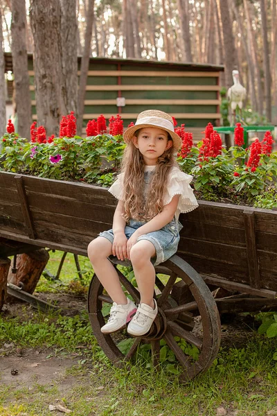 Preteen girl sitting on wheel of rustic wooden cart used as flower bed in city park — Stockfoto