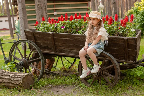 Расстроенная девочка-подросток, сидящая на колесе старинной деревянной тележки, украшенной как клумба — стоковое фото