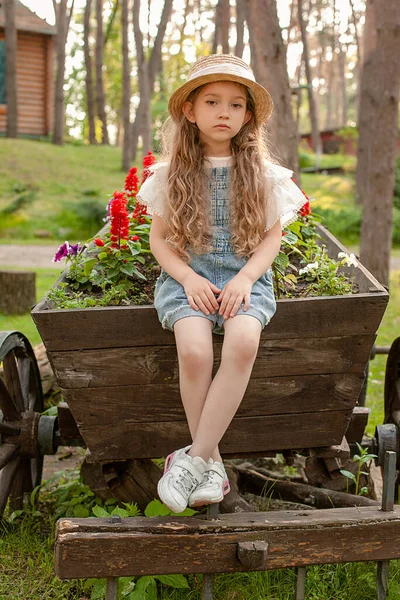 Сумна дівчинка сидить на старому дерев'яному візку, який використовується як клумба в літньому парку — стокове фото