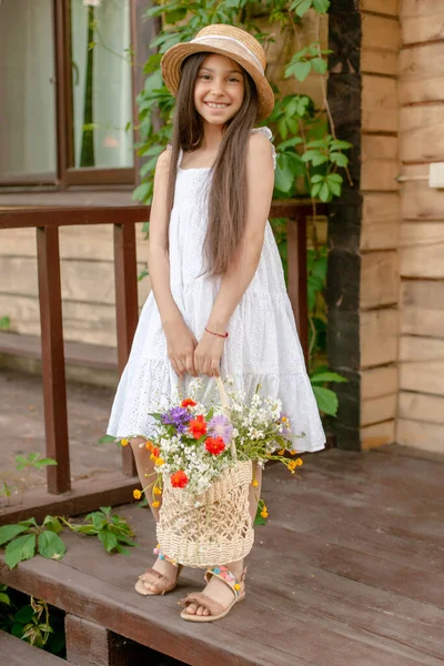 Gai preteen fille debout sur le seuil de chalet rural avec panier de fleurs sauvages — Photo
