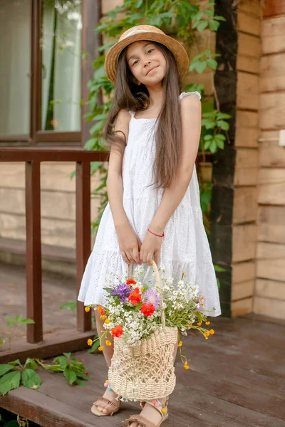 Glimlachende tiener brunette met mand van wilde bloemen staan op de drempel van het platteland huis — Stockfoto