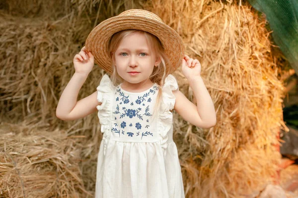 Linda niña rubia en vestido ligero con bordado y sombrero de paja de pie en el pajar — Foto de Stock