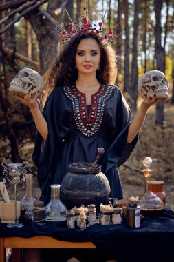 Siyah saçlı, uzun elbiseli, uzun kıvırcık saçlı bir cadı. Çam ormanında poz vermek. Kafataslarını tutmak. Büyü, büyü ve büyücülük. Yakın plan..