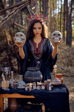 Siyah saçlı, uzun elbiseli, uzun kıvırcık saçlı bir cadı. Çam ormanında poz vermek. Kafataslarını tutmak. Büyü, büyü ve büyücülük. Yakın plan..