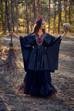 Siyah saçlı, uzun elbiseli, uzun saçlı bir cadı. Süpürge ve kafatası ile çam ormanında poz vermek. Büyü, büyü ve büyücülük. Tam uzunluk.