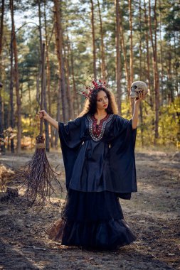 Siyah saçlı, uzun elbiseli, uzun saçlı bir cadı. Süpürge ve kafatası ile çam ormanında poz vermek. Büyü, büyü ve büyücülük. Tam uzunluk.