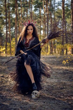 Siyah saçlı, uzun elbiseli, uzun saçlı bir cadı. Süpürge ve kafatası ile çam ormanında poz vermek. Büyü, büyü ve büyücülük. Tam uzunluk.