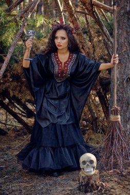 Siyah saçlı, uzun elbiseli, uzun saçlı bir cadı. Süpürge ve kafatası ile çam ormanında poz vermek. Büyü, büyü ve büyücülük. Tam uzunluk.