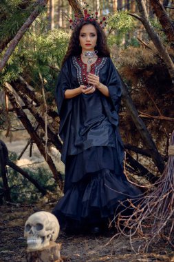 Siyah saçlı, uzun elbiseli, uzun saçlı bir cadı. Süpürge ve kafatası ile çam ormanında poz vermek. Büyü, büyü ve büyücülük. Tam uzunluk.