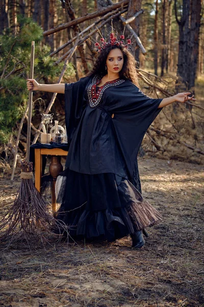 Boszorkány fekete, hosszú ruhában, vörös koronával a hosszú hajában. Seprűvel pózolok a fenyőerdőben. Varázslat, varázslat és boszorkányság. Teljes hosszúságú portré. — Stock Fotó