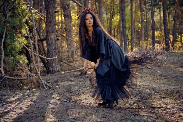 Pretty, wicked, long-haired sorceress in a black, long embroidered dress. There is large red crown in her brown, curly hair. She is posing sitting on her broom in a pine forest. Spells, magic and