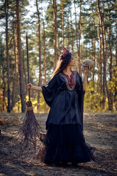 Häxa i svart, lång klänning, med röd krona i hennes långa hår. Possar med kvast och skalle i tallskogen. Trollformler, magi och häxeri. Full längd. — Stockfoto