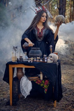 Siyah elbiseli, uzun elbiseli, saçları kırmızı taçlı bir cadı. Çam ormanında poz vermek, kafatası tutmak, iksir yapmak. Büyüler ve büyüler. Tam boy, duman..