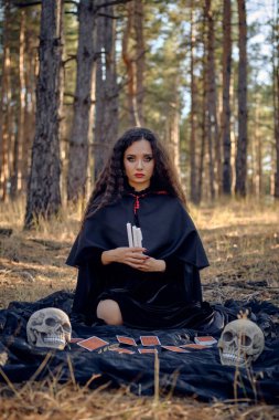 Pelerinli ve kapüşonlu siyah elbiseli cadı. Çam ormanında poz vermek. Üzerinde fal kartları ve kafatasları olan koyu bir battaniyenin üzerinde oturuyordu. Tam uzunluk.