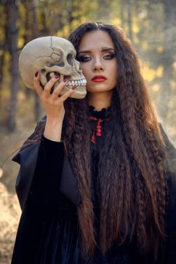 Siyah giysili, pelerinli ve başlıklı bir cadı. Dumanlı çam ormanında poz vermek. Kafatası tutuyorum. Büyü, büyü ve büyücülük. Yakın plan..