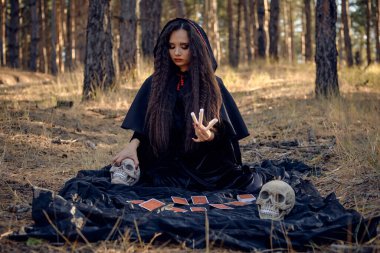 Pelerinli ve kapüşonlu siyah elbiseli cadı. Çam ormanında poz vermek. Üzerinde fal kartları ve kafatasları olan koyu bir battaniyenin üzerinde oturuyordu. Tam uzunluk.