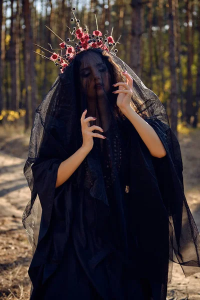 Häxa i svart, lång klänning, med röd krona i hennes långa, lockigt hår under en svart slöja. Possar i tallskog. Trollformler, magi och häxeri. Närbild. — Stockfoto