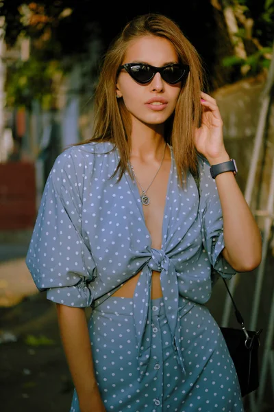 Blondes Mädchen in langem blauem Kleid, Sonnenbrille und kleiner schwarzer Handtasche auf der Schulter läuft allein durch die Stadt. Mode und Stil. Nahaufnahme. — Stockfoto