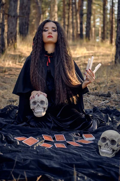 Hexe im schwarzen Kleid mit Umhang und Kapuze. Posieren im Kiefernwald. Auf einer dunklen Decke sitzend, mit Wahrsagerkarten und Totenköpfen darauf. Volle Länge. — Stockfoto