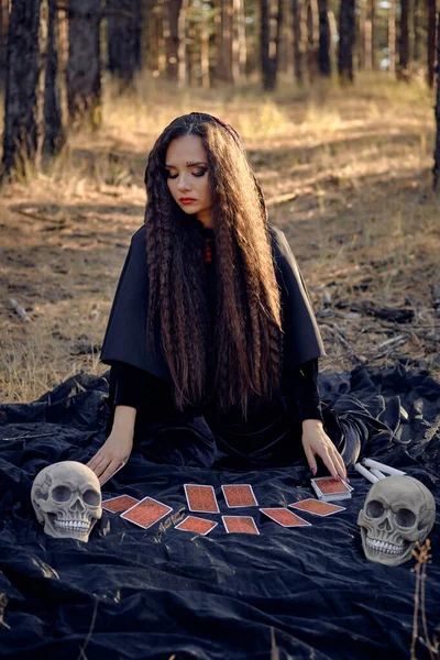 Hexe im schwarzen Kleid mit Umhang und Kapuze. Posieren im Kiefernwald. Auf einer dunklen Decke sitzend, mit Wahrsagerkarten und Totenköpfen darauf. Volle Länge. — Stockfoto