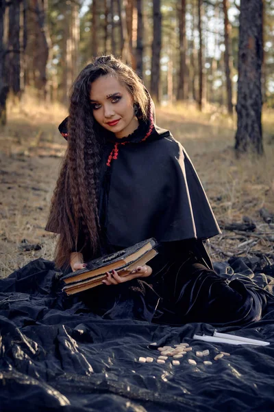 Sorcière en robe noire avec cape et capuchon. "Posing in pine forest". Assis sur une couverture sombre, des livres, des bougies et des runes. Longueur totale. — Photo