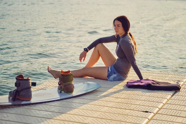 Brunette girl in a gray turtleneck and blue denim shorts is posing with her wakeboard on a pier of the coastal zone. — Stock Fotó