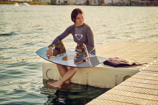Brunette girl in a gray turtleneck and blue denim shorts is posing with her wakeboard on a pier of the coastal zone. — Fotografia de Stock