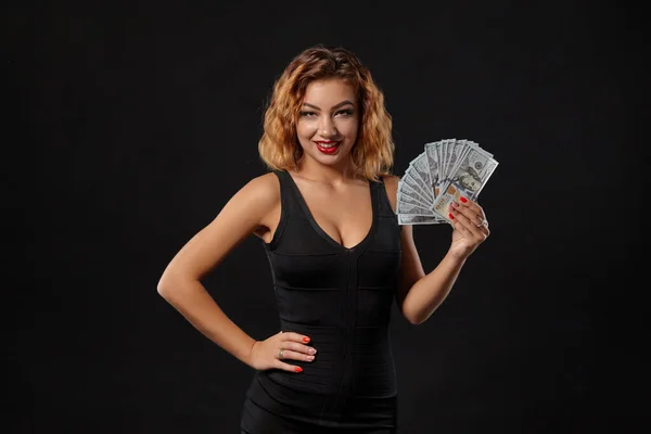 Imbirowa dziewczyna w ciemnej sukience, pozująca trzymając w rękach fankę studolarowych banknotów stojących na tle czarnego studia. Kasyno, poker. Zbliżenie. — Zdjęcie stockowe
