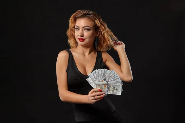 Imbirowa dziewczyna w ciemnej sukience, pozująca trzymając w rękach fankę studolarowych banknotów stojących na tle czarnego studia. Kasyno, poker. Zbliżenie. — Zdjęcie stockowe