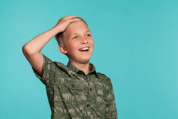 青いスタジオの背景にヤシのプリントがポーズをとっている緑色のシャツの中のブロンドの10代の少年のクローズアップ肖像。誠実な感情の概念. — ストック写真