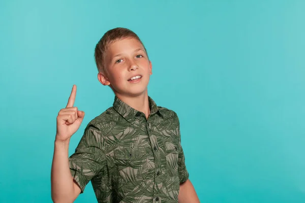 Close-up portrait of a blonde teenage boy in a green shirt with palm print posing against a blue studio background. Concept of sincere emotions. — Stockfoto