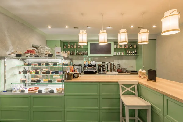 Interior of cozy coffeehouse in Provence style with green pastel furniture fronts on counter, decorative hanging fabric lampshades in shape of milk cans and glass showcase with various tempting sweets