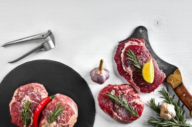 Siyah tahtada taze çiğ biftek, sarımsak, zeytinyağı, biber, tuz ve biberiye: bonfile, Striploin, kaburga gözü ve beyaz arka planda bir mutfak baltası. Üst Manzara. Uzayı kopyala Durgun hayat