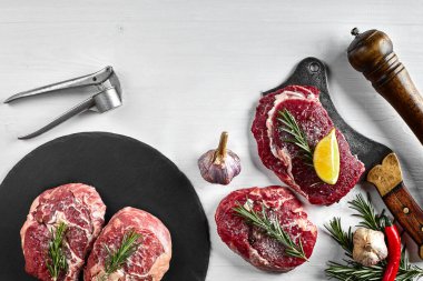 Siyah tahtada taze çiğ biftek, sarımsak, zeytinyağı, biber, tuz ve biberiye: bonfile, Striploin, kaburga gözü ve beyaz arka planda bir mutfak baltası. Üst Manzara. Uzayı kopyala Durgun hayat