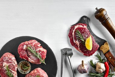 Siyah tahtada taze çiğ biftek, sarımsak, zeytinyağı, biber, tuz ve biberiye: bonfile, Striploin, kaburga gözü ve beyaz arka planda bir mutfak baltası. Üst Manzara. Uzayı kopyala Durgun hayat