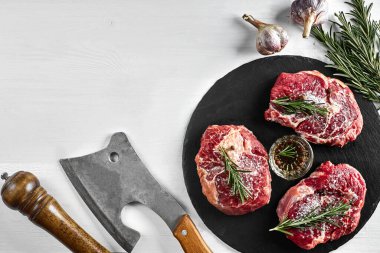 Siyah tahtada taze çiğ biftek, sarımsak, zeytinyağı, biber, tuz ve biberiye: bonfile, Striploin, kaburga gözü ve beyaz arka planda bir mutfak baltası. Üst Manzara. Uzayı kopyala Durgun hayat