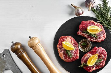 Siyah tahtada taze çiğ biftek, sarımsak, zeytinyağı, biber, tuz ve biberiye: bonfile, Striploin, kaburga gözü ve beyaz arka planda bir mutfak baltası. Üst Manzara. Uzayı kopyala Durgun hayat