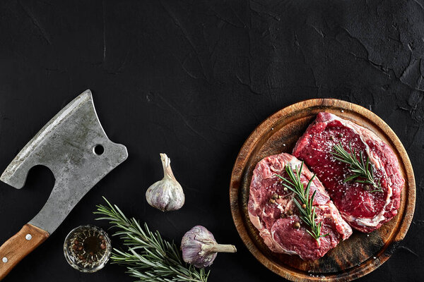 Fresh raw meat. Beef. Piece of beef steak tenderloin, with ax for cutting and chopping meat, spices were cooking - rosemary, pepper, salt, garlic. On old wooden board on stone black table. Top view