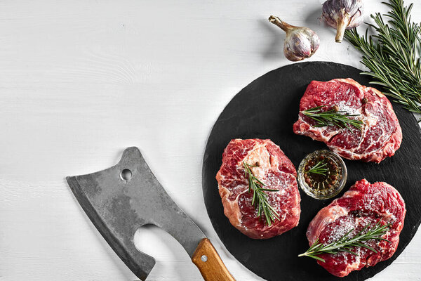 Fresh raw beef steaks with herbs, garlic, olive oil, pepper, salt and rosemary on black board: Tenderloin, Striploin, Rib Eye and a kitchen ax on white background. Top view. Copy space. Still life