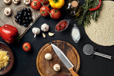 Tahta kesme tahtası ve geleneksel pizza hazırlama malzemeleri: peynir, domates sosu, mantar, zeytinyağı, biber, baharatlar. Siyah desenli masa arkaplanı. Üst Manzara. Durgun hayat. Boşluğu kopyala