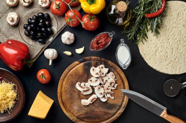 Tahta kesme tahtası ve geleneksel pizza hazırlama malzemeleri: peynir, domates sosu, mantar, zeytinyağı, biber, baharatlar. Siyah desenli masa arkaplanı. Üst Manzara. Durgun hayat. Boşluğu kopyala