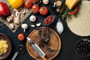 Tahta kesme tahtası ve geleneksel pizza hazırlama malzemeleri: peynir, domates sosu, zeytin, zeytin yağı, biber, baharatlar. Siyah desenli masa arkaplanı. Üst Manzara. Durgun hayat. Uzayı kopyala Düz
