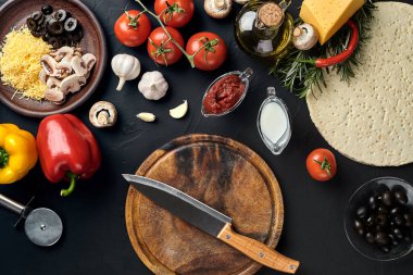 Tahta kesme tahtası ve geleneksel pizza hazırlama malzemeleri: peynir, domates sosu, fesleğen, zeytinyağı, biber, baharatlar. Siyah desenli masa arkaplanı. Üst Manzara. Durgun hayat. Uzayı kopyala Düz