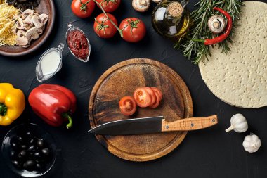 Tahta kesme tahtası ve geleneksel pizza hazırlama malzemeleri: peynir, domates, sos, zeytin, zeytin yağı, biber, baharatlar. Siyah desenli masa arkaplanı. Üst Manzara. Durgun hayat. Uzayı kopyala Düz
