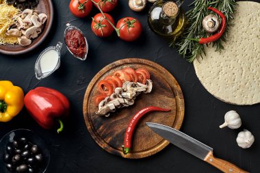 Tahta kesme tahtası ve geleneksel pizza hazırlama malzemeleri: peynir, domates, sos, mantar, zeytinyağı, biber, baharatlar. Siyah desenli masa arkaplanı. Üst Manzara. Durgun hayat. Boşluğu kopyala