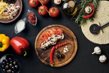 Tahta kesme tahtası ve geleneksel pizza hazırlama malzemeleri: peynir, domates, sos, mantar, zeytinyağı, biber, baharatlar. Siyah desenli masa arkaplanı. Üst Manzara. Durgun hayat. Boşluğu kopyala