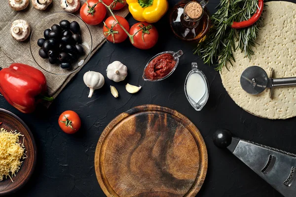 Tahta kesme tahtası ve geleneksel pizza hazırlama malzemeleri: peynir, domates sosu, fesleğen, zeytinyağı, biber, baharatlar. Siyah desenli masa arkaplanı. Üst Manzara. Durgun hayat. Uzayı kopyala Düz