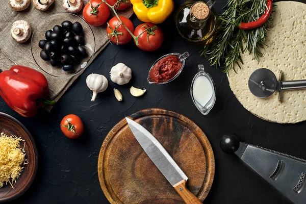 Tahta kesme tahtası ve geleneksel pizza hazırlama malzemeleri: peynir, domates sosu, fesleğen, zeytinyağı, biber, baharatlar. Siyah desenli masa arkaplanı. Üst Manzara. Durgun hayat. Uzayı kopyala Düz
